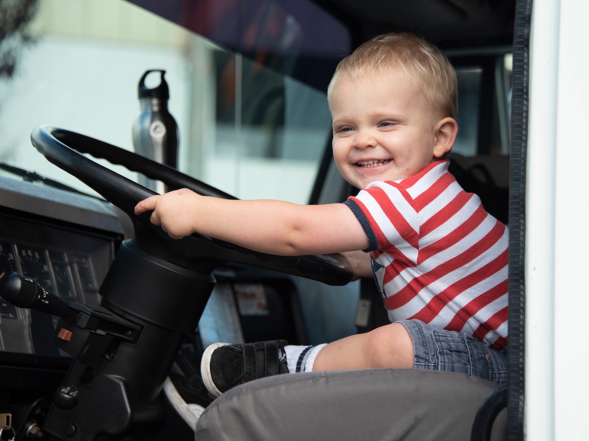 A young boy in a red striped shirt smiles while holding a big steering wheel in the cab of truck.
