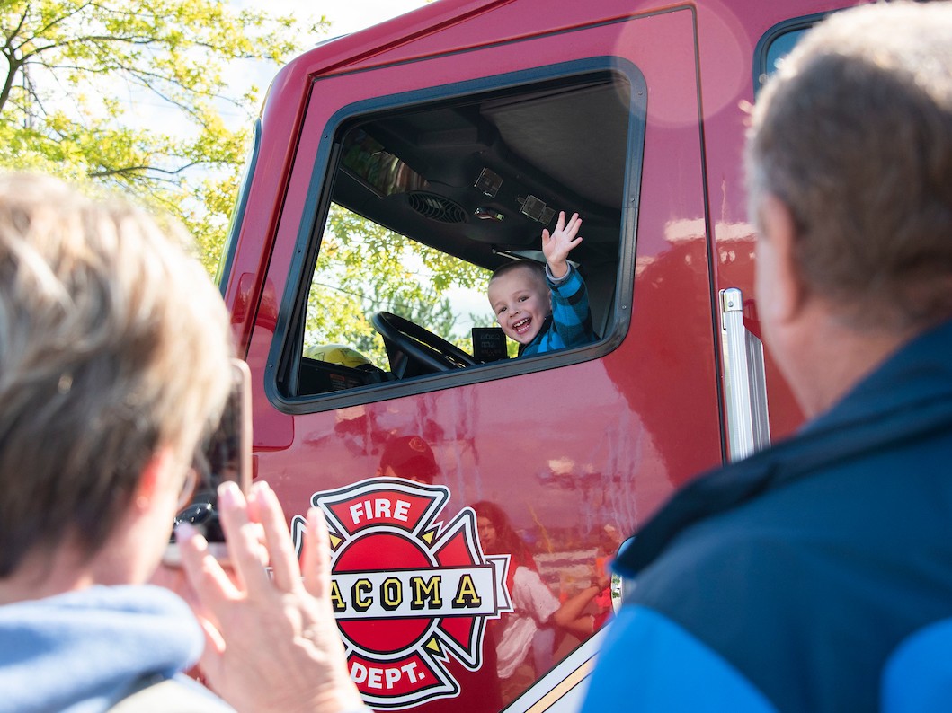 A small boy waves at caregivers while sitting in the cab of a firetruck. 