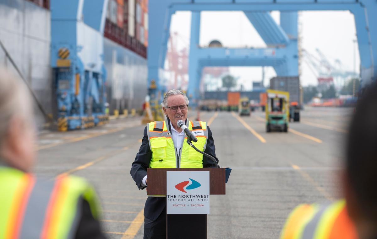 An older man in a reflective vest speaks at a podium on a port terminal.