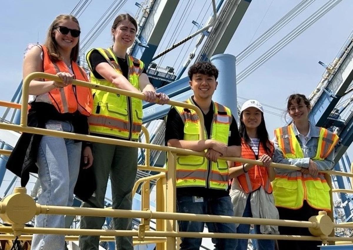 A group of college students in high visibility vests smile while standing on the deck of a boat.