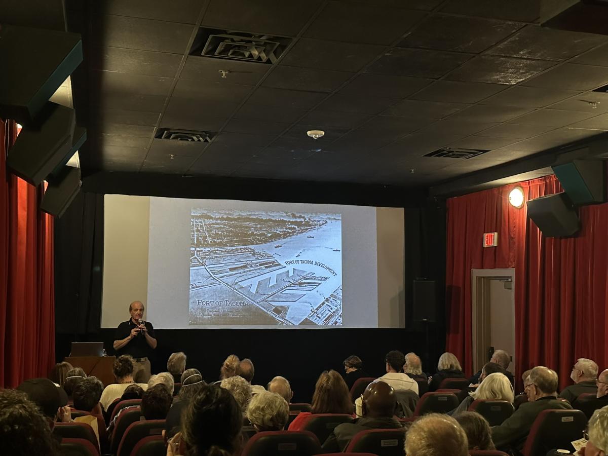 A man presents in the front of a theater filled with people. 